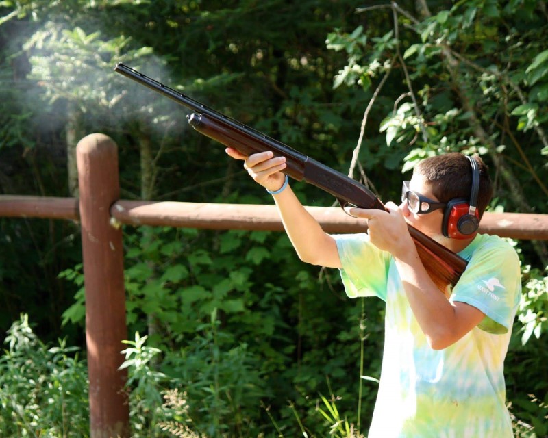 A Scout in safety glasses and earmuffs shooting a shotgun. The end of the gun is surrounded a puff of smoke from firing the gun