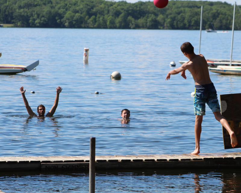 A Scout throwing a basketball back to two Scouts swimming in the lake