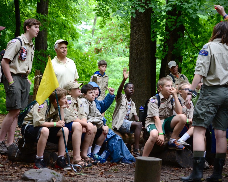 Several younger Scouts raise their hands to ask questions as two staff give a presentation to a large group of Scouts