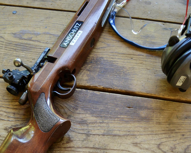 A rifle, pair of safety glasses, and earmuffs on a wooden plank table