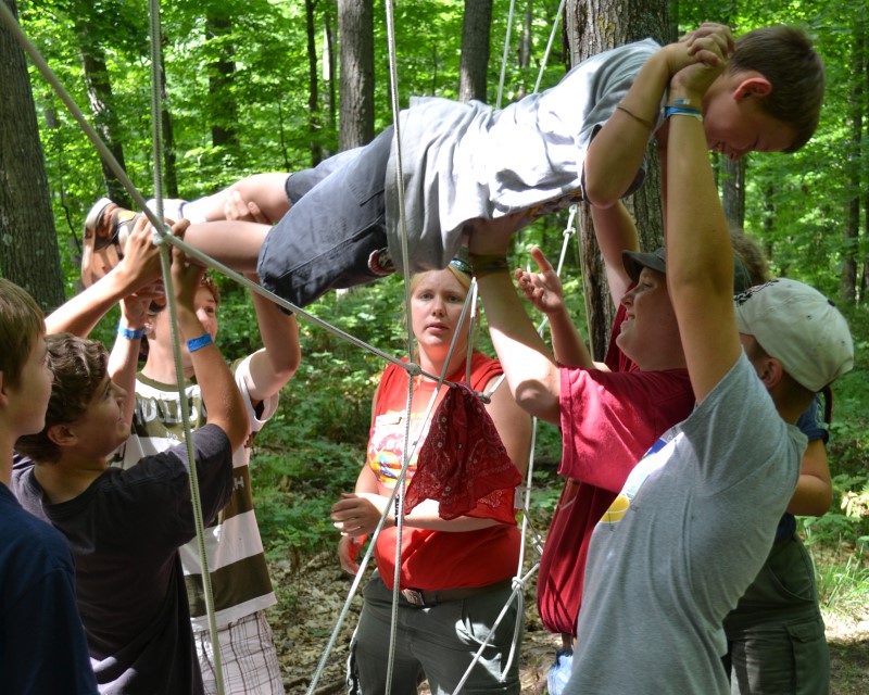 A group of Scouts carrying a Scout through a hole in a wall of rope